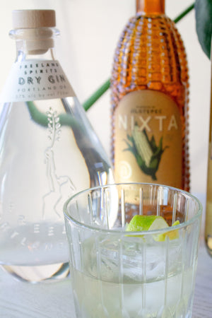 Bottles of Freeland Spirits Dry Gin, Nixta, and Mezcal on a white table surrounded by monsterra plant fronds. Cocktail in fron with a lime twist.