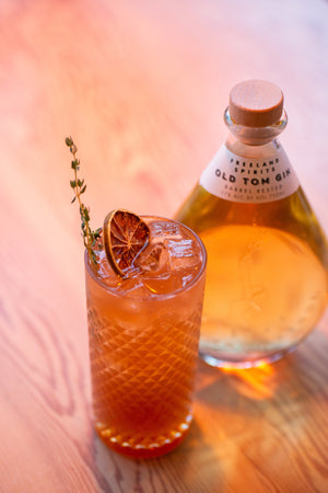 Classic Collins gin cocktail garnished with dried citrus and thyme, next to bottle of Freeland Old Tom Gin