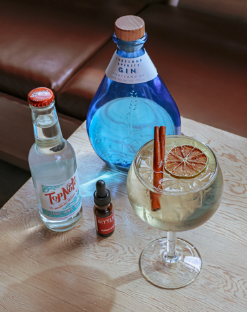 Freeland Fall Gin & Tonic cocktail garnished with a cinnamon stick, next to bottle of Freeland Gin, Top Note Classic Tonic, and PDX Bitters