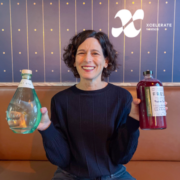 Abby Guyer, Executive Director of Xcelerate Women in Portland, OR smiling at Freeland Spirits tasting room & distillery while holding bottle of Forest Gin and Passion Potion fresh cocktail mixer