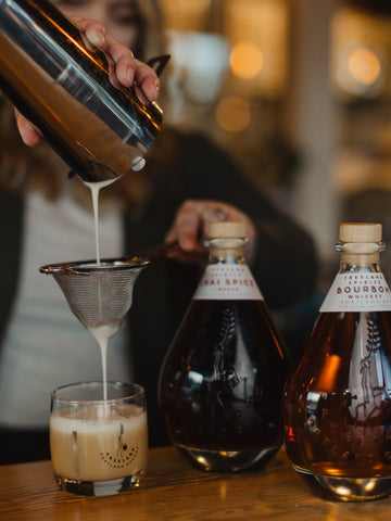 Cocktail being poured into a Freeland Spirits rocks glass, next to bottles of Freeland Bourbon and Smith Tea Collaboration Chai Spice Liqueur 